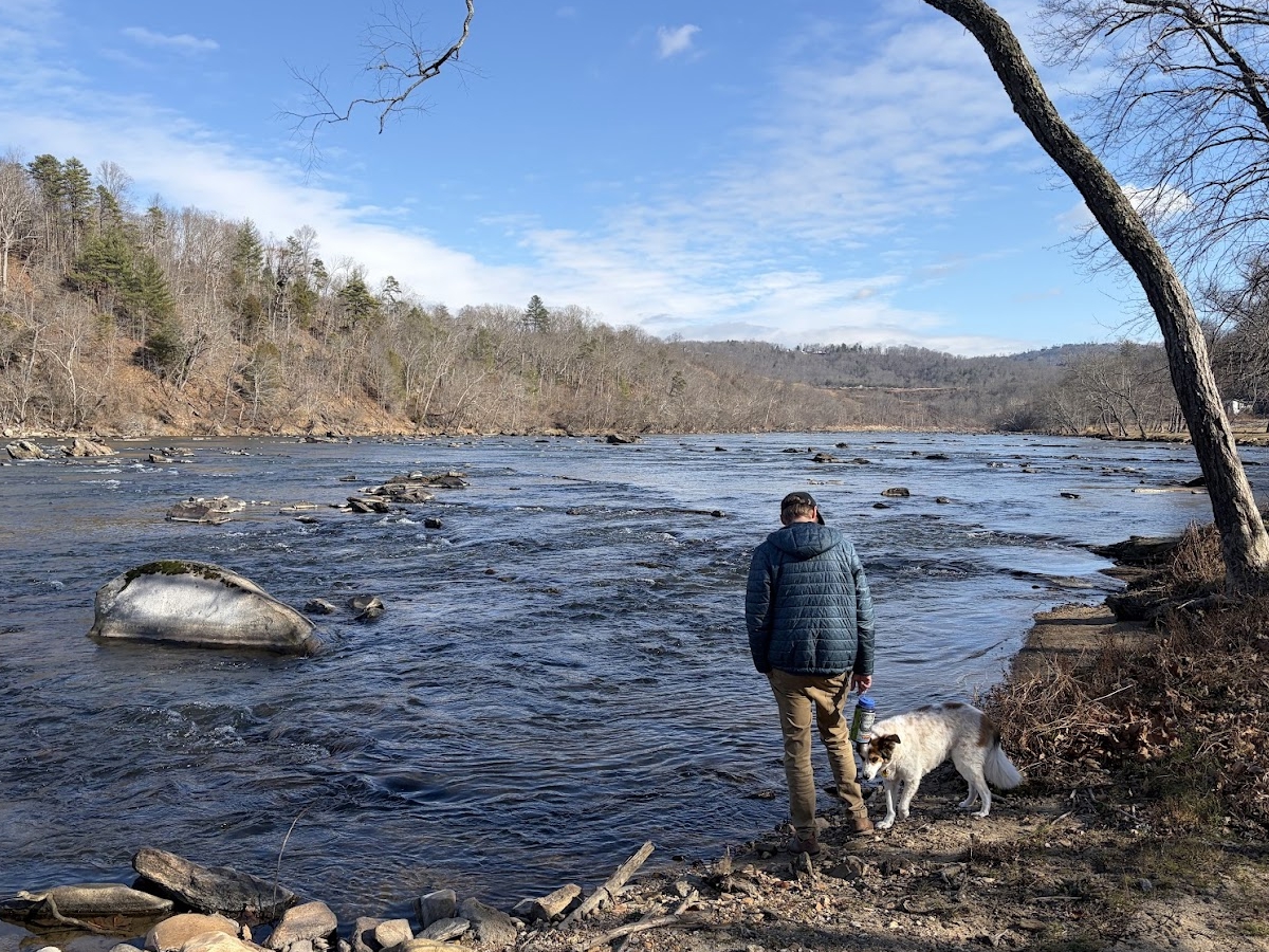 Scott stands on the river’s edge holding a water bottle while Pixie, a white-and-brown dog, sniffs the ground; a broad, rocky river flows past leafless trees under a bright winter sky.